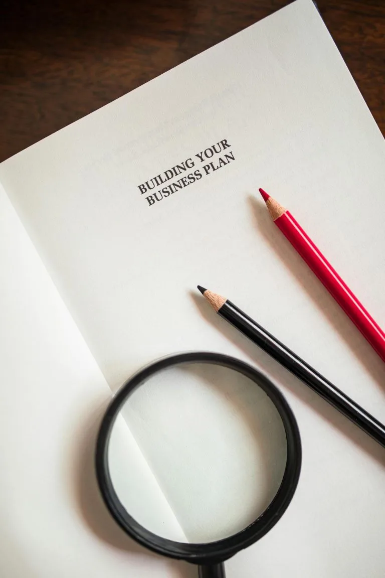 Top view of a business plan document with a magnifying glass and colored pencils.