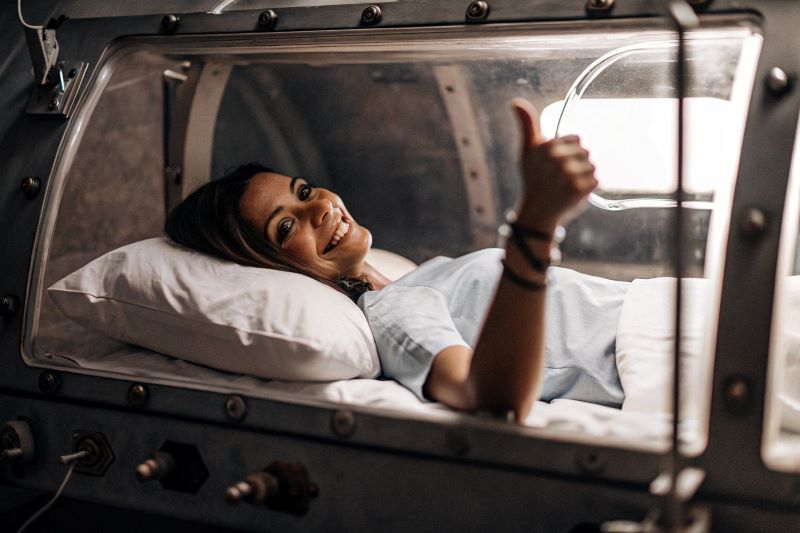 A woman lying in a hyperbaric oxygen therapy chamber with a thumbs up